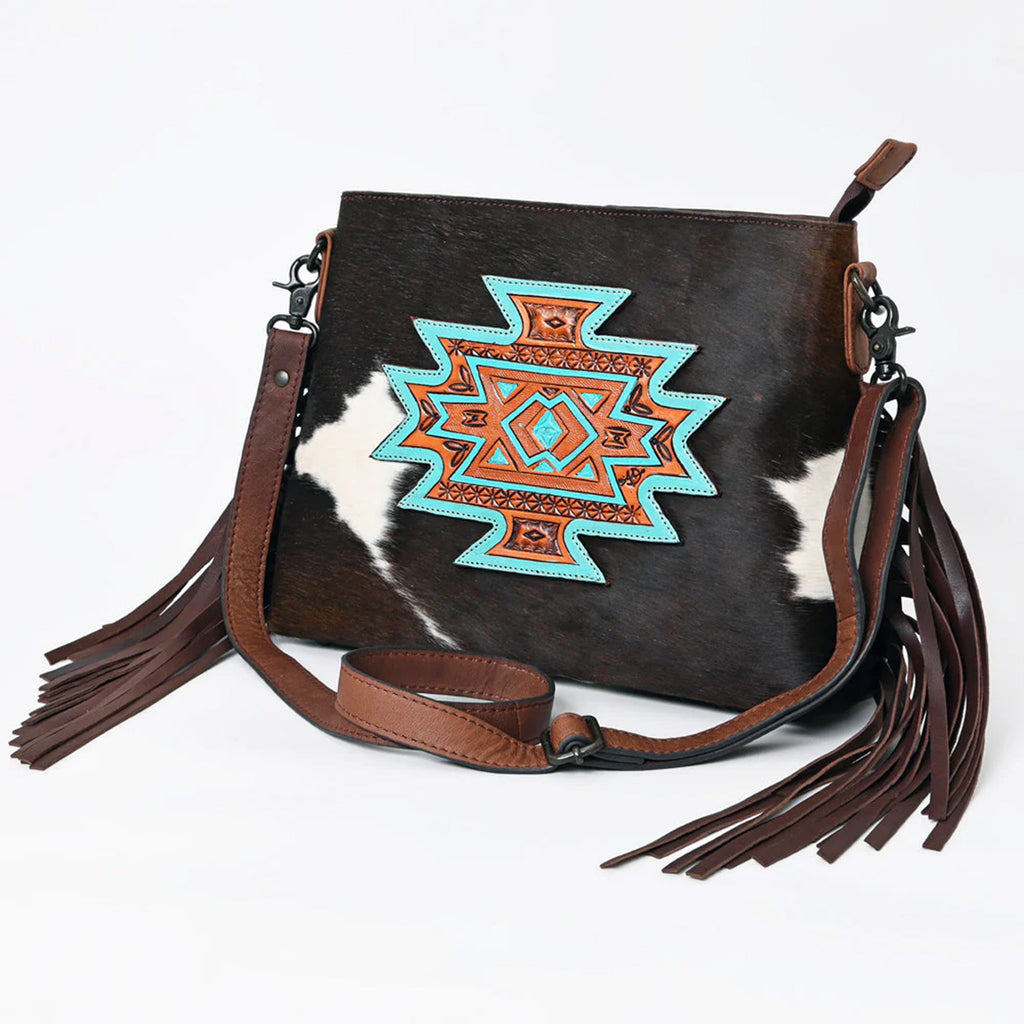 Black and white cowhide purse with large turquoise and brown tooled leather patch, fringe on the sides, and shoulder strap.