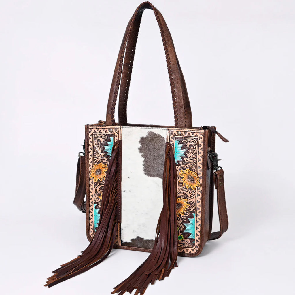 Leather tooled bag with the middle being black and white cowhide and the sides having leather tooling with sunflowers and Aztec designs. Bag also features fringes on the front, two handles, and a long shoulder strap. 
