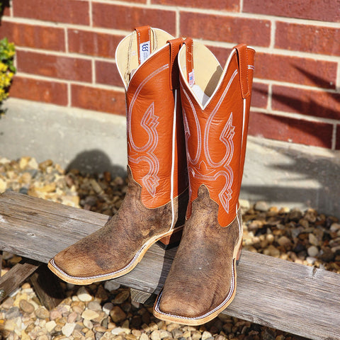 Pair of brown and orange cowboy boots on a wooden bench with a brick wall background.