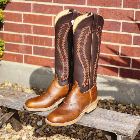 Pair of brown cowboy boots with intricate designs on a wooden bench against a brick wall.