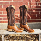 Pair of brown cowboy boots with intricate designs on a wooden bench against a brick wall.