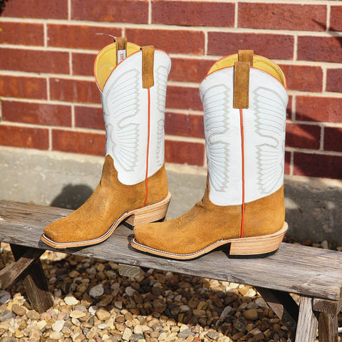 Pair of tan cowboy boots with white tops on a wooden bench against a brick wall.