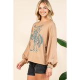 Female model shown at an angled side‑front pose wearing the mocha long sleeve shirt, displaying the turquoise bronc rider graphic.