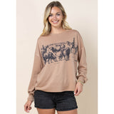 Female model wearing a camel long sleeve shirt featuring a herding‑cattle Western graphic, shown to highlight the fit, sleeve detail, and ranch‑inspired design.