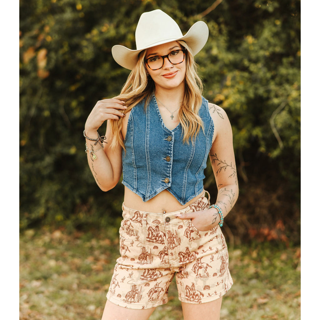 Woman wearing a denim vest and western patterned shorts with a cowboy hat.