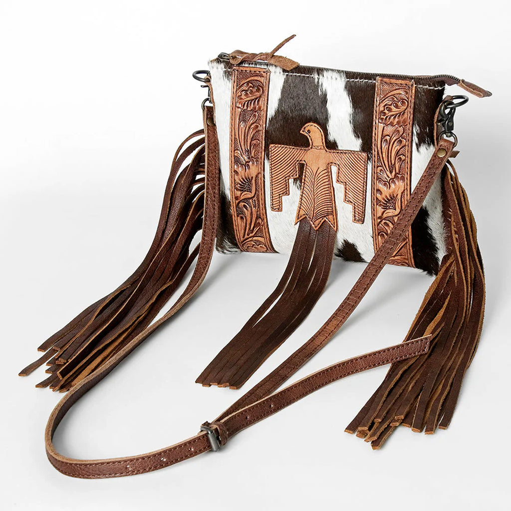 Black and white cowhide purse with adjustable leather shoulder strap, tooled leather on the sides, brown fringe on the sides. and a large thunderbird on the front. 