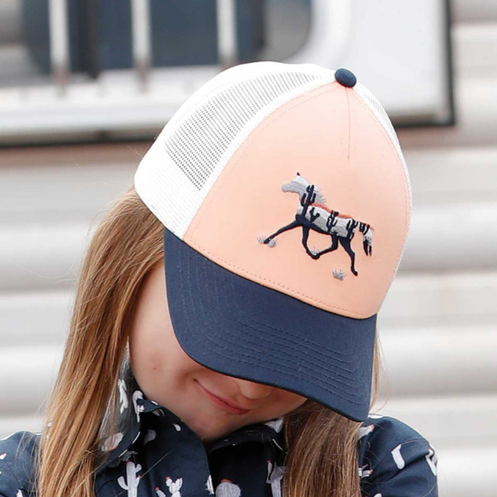 Girl model wearing hat with head tilted down. Hat has a peach colored front with a stitched desert horse patch, white mesh backing, and navy blue bill. 