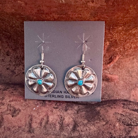 Silver earrings with turquoise stones on a gray card against a red-brown stone background