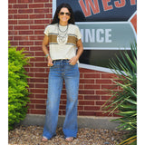 Woman in a striped shirt and blue jeans standing in front of a brick wall with a sign.