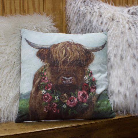 Small throw pillow of a painted highland cow with a garland of pink roses around its neck, pillow is sitting between two other white furry pillows. 
