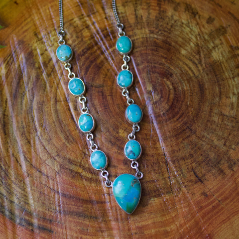 Flat lay of silver and turquoise necklace. It has nine stones, one large upside down teardrop stone in the middle and four smaller oval stones on each side. 