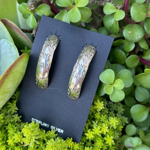 Hoop earrings with an engraved tribal design, on a black card with 'STERLING SILVER' text, surrounded by green plants.