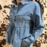 Model is wearing long sleeve denim pearl snap with white stitched loop detailing. Shirt flares out under the chest.