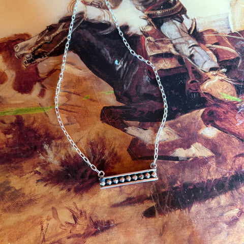 Silver bar necklace and chain with silver dots going horizontal with the bar, necklace is laying flat on a western designed table.