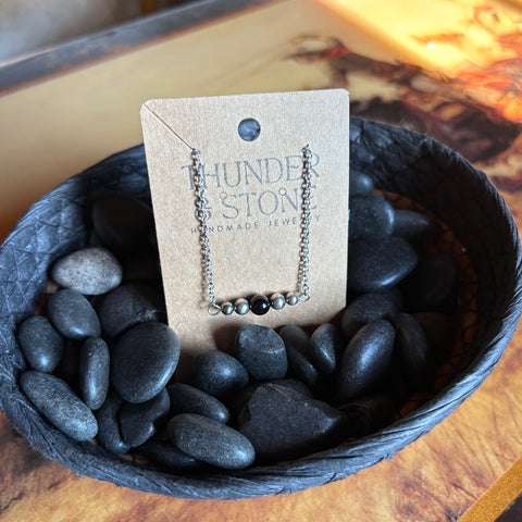 Navajo style pearl necklace with two Navajo style pearls on each side and one black onyx stone in the middle with a sterling silver chain, necklace is attached to a Thunder & Stone label and is sitting in a basket with black stones. 