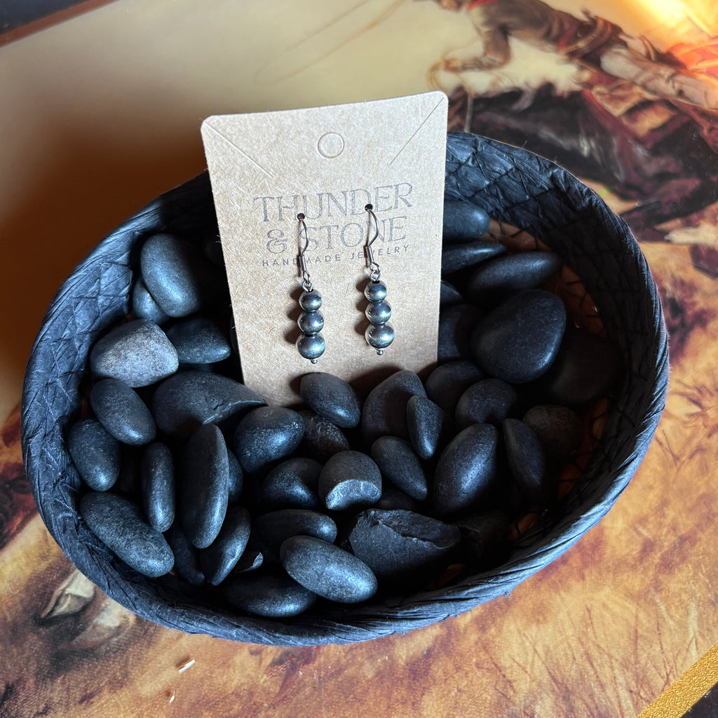 Navajo style pearl earrings with three large pearls stacked on each other, earrings are attached to a Thunder & Stone label and sitting in a basket of black stones. 