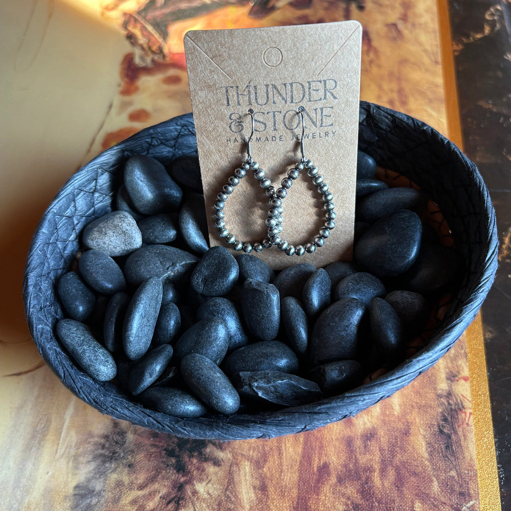 4mm Navajo pearl earrings in a teardrop hoop shape. Earrings are attached to a Thunder & Stone label and sitting in a basket of black stones. 
