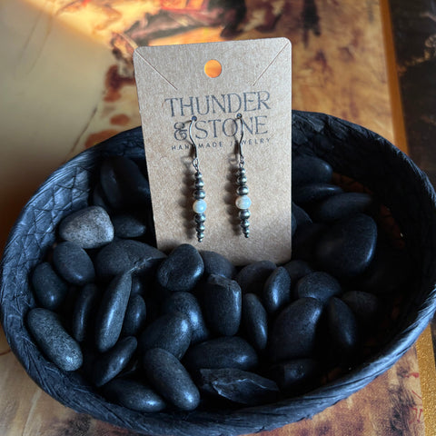 Graduated straight string of Navajo style pearls with a white pearl in the middle, earrings are attached to a Thunder & Stone label and sitting in a basket of black stones.