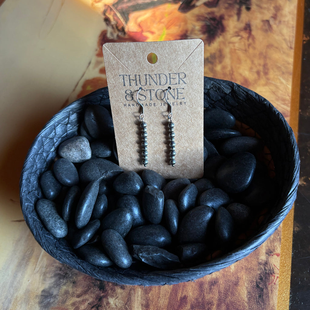 Straight string of nine small Navajo pearl earrings, earrings are attached to a Thunder & Stone label, sitting in a basket of black stones.