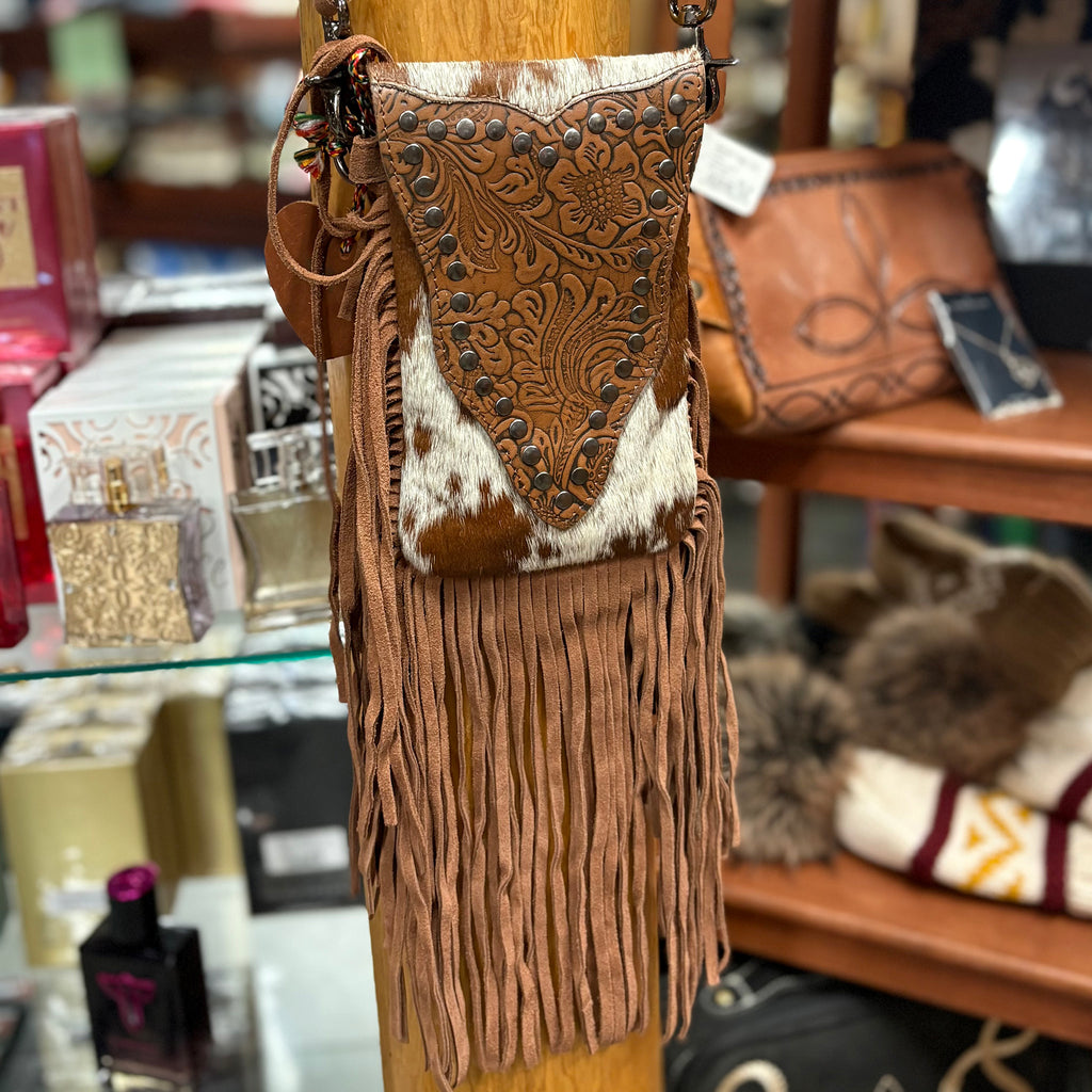 Small travel purse with brown and white cowhide, tooled leather on the flap, metal studs, and long brown leather fringe.