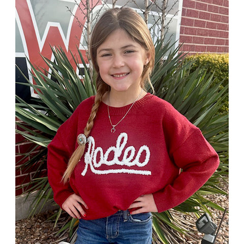 Young girl wearing a red sweater with 'Rodeo' text, standing outdoors.