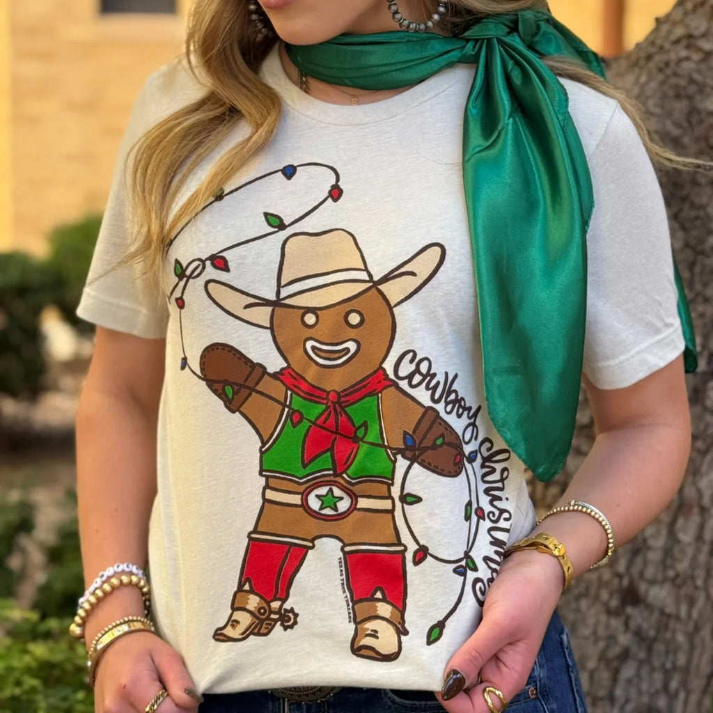 Cream colored tee with a gingerbread man in a cowboys outfit swinging a Christmas light rope and cursive writing that says "Cowboy Christmas"