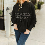 Model is wearing a black long sleeve button up with pearl snaps. Button up flares out under the chest and has two white stitched loop designs and one straight line between them.