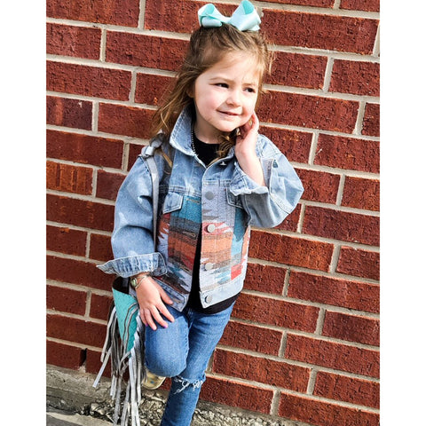 A little girl wearing the Girls Denim Aztec Jacket, posed against a brick building.