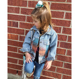 A little girl wearing the Girls Denim Aztec Jacket, looking back at brick building.