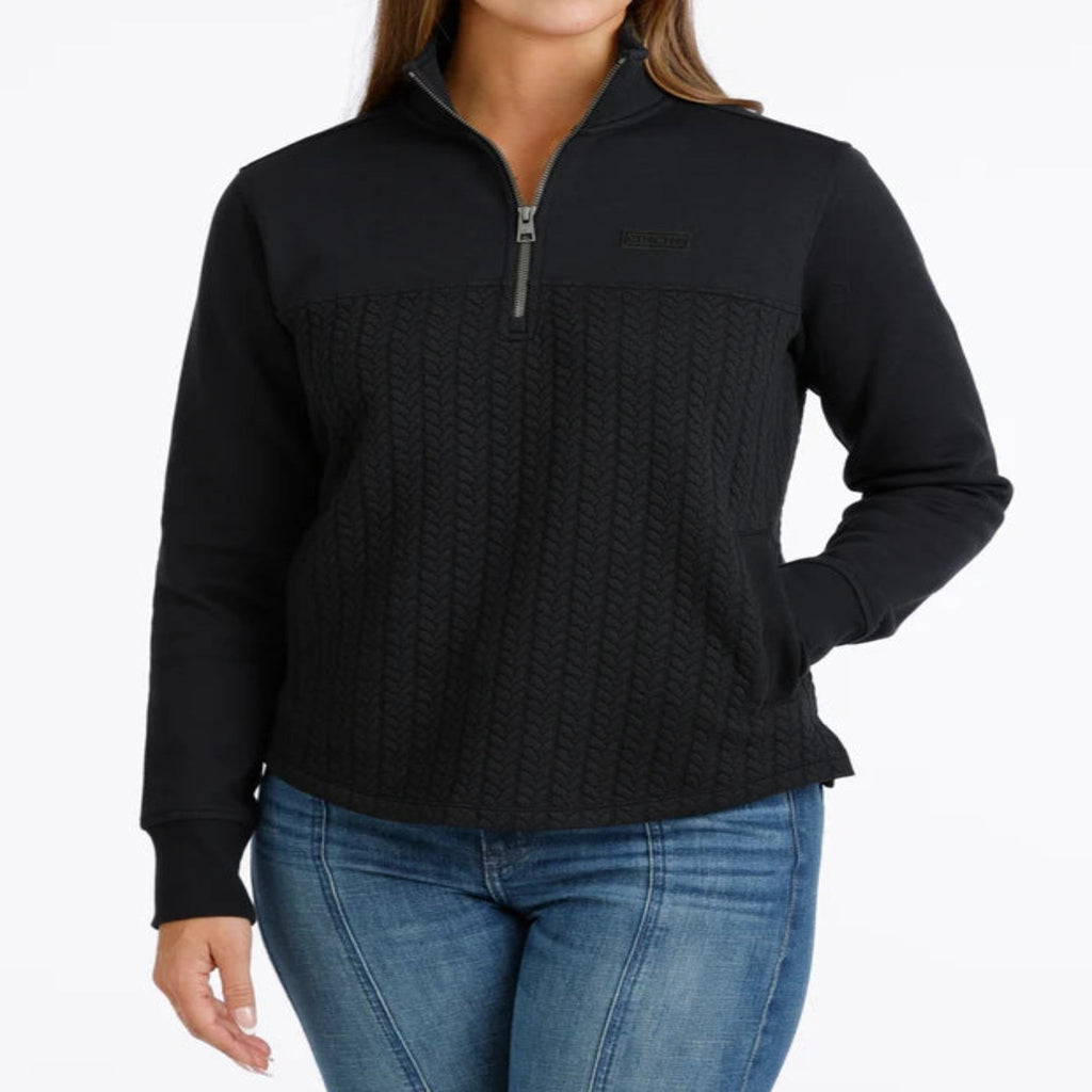 Woman wearing a black quarter-zip pullover with a textured cable-knit front, stand collar, side pockets, and subtle CINCH logo.