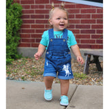 Child wearing blue denim overalls with a horse design in front of a brick wall.