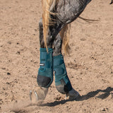 Horse wearing teal protective leg wraps on a dirt ground