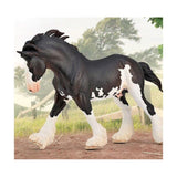 Model horse with black and white coat standing on a dirt path with greenery