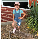 Child wearing a pink cap and denim overalls sitting on a bench in front of a brick building.