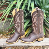 Pair of brown cowboy boots with decorative patterns on a wooden surface with green plants in the background.