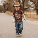 Boy model wearing layered long sleeve shirt with grey long sleeves and brown t-shirt with large rodeo/cowboy motif on the front. 